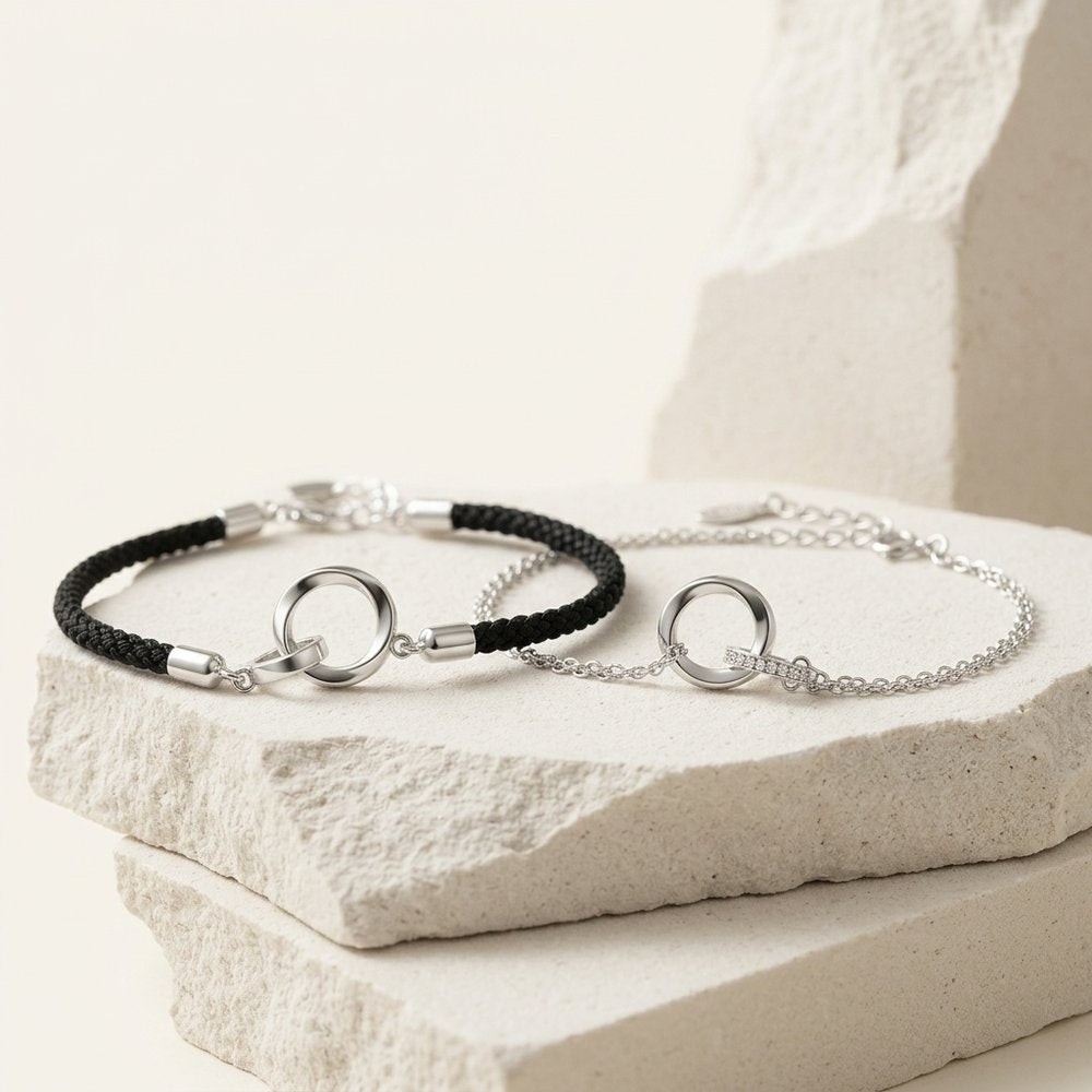 Deux bracelets anneaux entrelacés, chaîne argentée et cordon noir, posés sur un support minéral ivoire