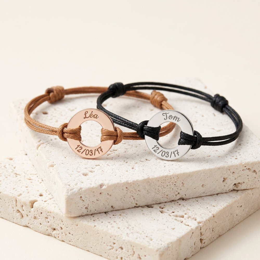 Deux bracelets couple en cordon avec anneau gravé en acier inoxydable, posés sur un support minéral ivoire