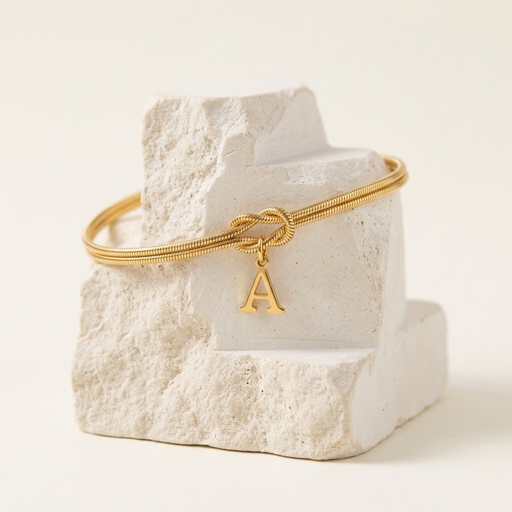 Bracelet noeud initial doré avec breloque lettre posé sur un support minéral ivoire