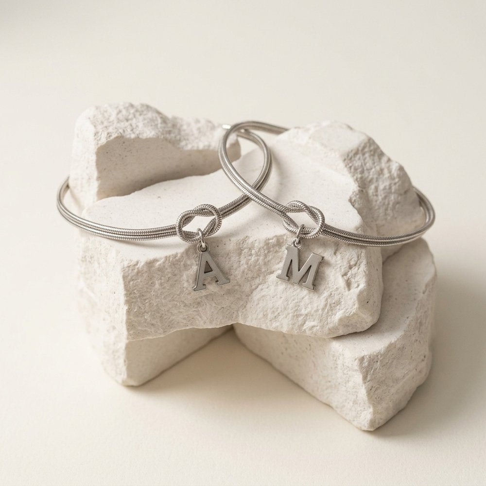 Deux bracelets noeud couple en acier inoxydable avec initiale, posés sur un support minéral ivoire