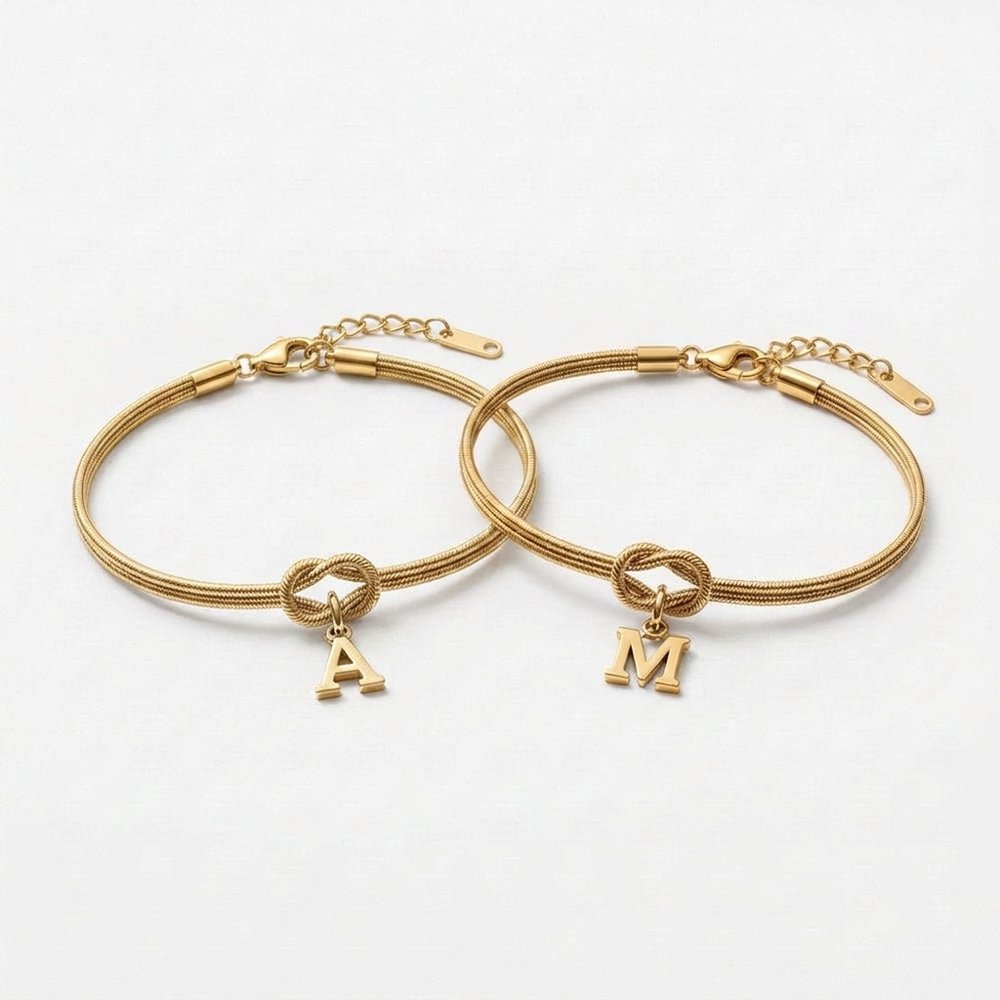 Duo de bracelet noeud doré : deux chaînes serpent or avec nœud d'amour et initiales A et M, sur fond neutre